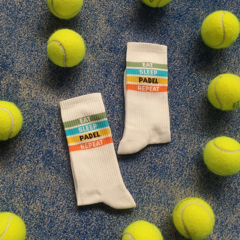 May include: Two pairs of white socks with colorful stripes and the text "Eat Sleep Padel Repeat" printed on them. The socks are surrounded by yellow tennis balls.