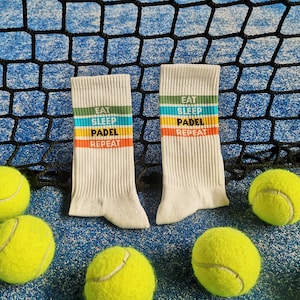 May include: Two white socks with colorful stripes and the text "Eat Sleep Padel Repeat" on them. The socks are lying on a blue surface with yellow tennis balls.