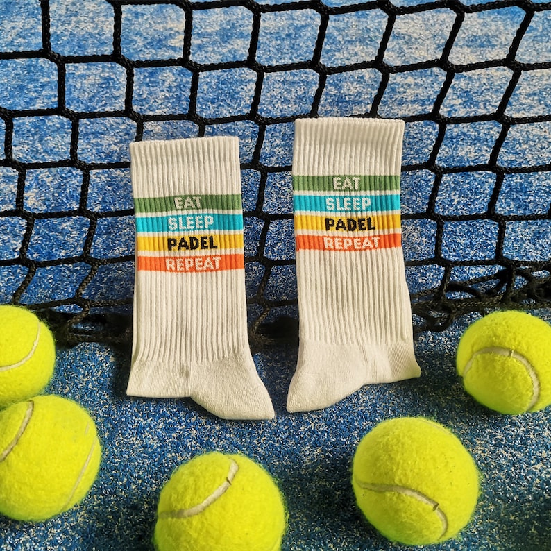 May include: Two white socks with colorful stripes and the text "Eat Sleep Padel Repeat" on them. The socks are lying on a blue surface with yellow tennis balls.