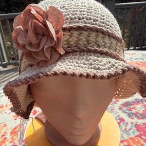 May include: A cream-colored crochet hat with a wide brim and a decorative band. A large, light-brown fabric flower adorns the side. The hat is displayed on a mannequin head.
