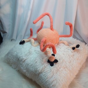 Peut inclure: Une peluche de pieuvre en crochet rose et blanche avec des yeux noirs, posée sur un coussin blanc moelleux.