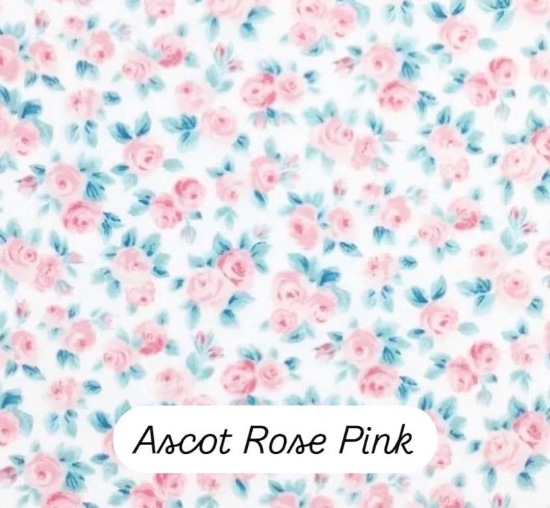 May include: A fabric with a repeating floral pattern of pink roses and blue-green leaves on a white background. The text "Ascot Rose Pink" is displayed in a white rectangle at the bottom of the image.