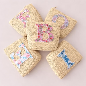 May include: Five beige woven straw pouches arranged in a circular pattern. Each pouch features a large, fabric-appliqued letter on the front: E, O, B, E, and I. The letters are decorated with various floral and strawberry patterns in pink, blue, and red.
