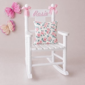 May include: A white wooden rocking chair featuring the name Rosie in pink script on the backrest. The chair is adorned with two pink gingham bows on the top posts and includes a square cushion with a pink rose and green leaf floral pattern.