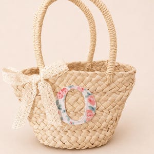 May include: A woven straw basket with two rounded handles, featuring a beige color. The front is decorated with a white lace bow tied on the left side and a large, fabric-stitched letter O with a pink and green floral pattern centered on the body.