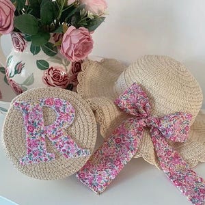 May include: A straw sun hat with a floral trim and a matching floral ribbon tied in a bow. The hat is paired with a straw coaster with a floral fabric letter R.