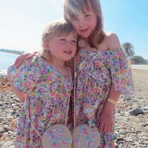 May include: Two young girls wearing matching floral print dresses and round straw purses with the letters "R" and "N". The dresses have off-the-shoulder sleeves and a colorful floral pattern. The girls are standing on a beach.