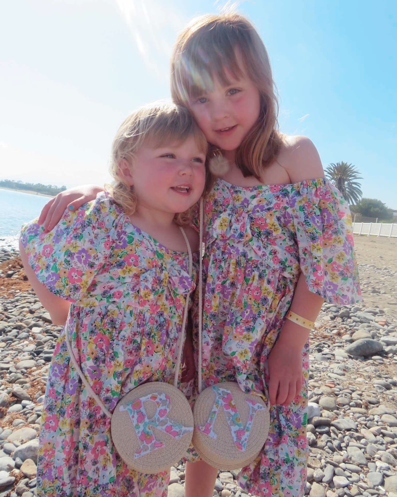 May include: Two young girls wearing matching floral print dresses and round straw purses with the letters "R" and "N". The dresses have off-the-shoulder sleeves and a colorful floral pattern. The girls are standing on a beach.