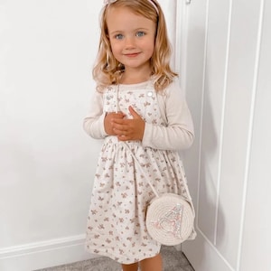 May include: A young girl wearing a white floral dress with a white headband and a white straw bag with a pink and silver letter A on it.