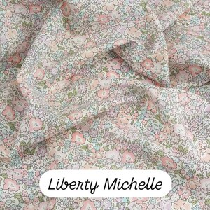 May include: Close-up of Liberty Michelle fabric. The fabric features a delicate floral pattern in shades of pink, peach, lavender, and light blue on a cream background. The fabric is tightly woven and appears soft, with a slight sheen. The text "Liberty Michelle" is displayed in a white oval.