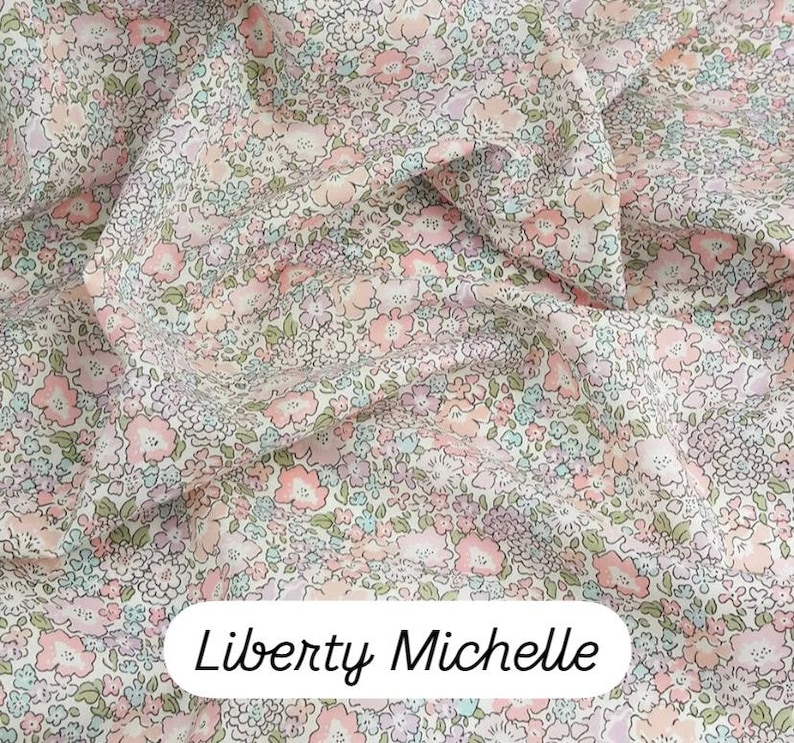 May include: Close-up of Liberty Michelle fabric. The fabric features a delicate floral pattern in shades of pink, peach, lavender, and light blue on a cream background. The fabric is tightly woven and appears soft, with a slight sheen. The text "Liberty Michelle" is displayed in a white oval.