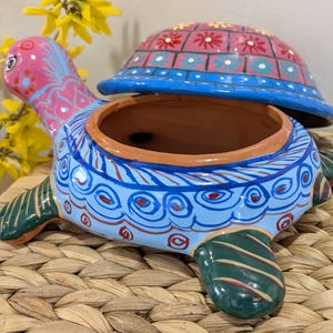 May include: Hand-painted ceramic turtle-shaped container with a removable lid. The turtle has a pink head, blue body, and green flippers. The lid is pink with a floral pattern. The container is on a woven surface.