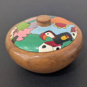 Peut inclure: Petite boîte à bijoux ronde en bois avec couvercle. Le couvercle est peint d'un motif coloré représentant un toucan, des maisons et du feuillage. La boîte est de couleur marron chaud, avec un petit bouton en bois.