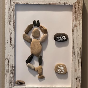 May include: Framed artwork featuring a yoga pose created from various-sized stones. The stones are arranged on a white background within a distressed white wooden frame. Additional stones display a lotus flower and an Om symbol.