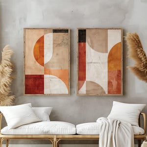 May include: Two framed abstract art prints. Each print features geometric shapes in shades of orange, red, beige, and white. The artwork is displayed above a wicker bench with white cushions and pillows.