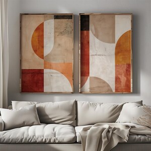 May include: Two framed abstract art prints. Each print features a geometric design with orange, red, white, and beige shapes. The artwork has a textured, vintage appearance, with a wooden frame. The prints are displayed above a white sofa.