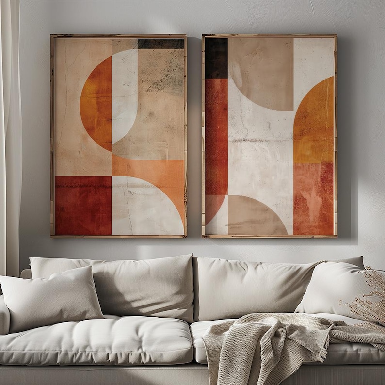 May include: Two framed abstract art prints. Each print features a geometric design with orange, red, white, and beige shapes. The artwork has a textured, vintage appearance, with a wooden frame. The prints are displayed above a white sofa.