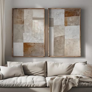 May include: Two abstract paintings in wooden frames, each featuring a geometric pattern of beige, brown, and white squares and rectangles. The paintings are hung on a light gray wall above a white sofa.