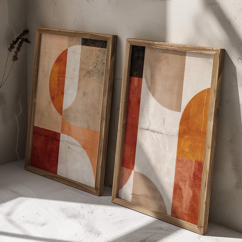 May include: Two framed abstract art prints. Each print features geometric shapes in shades of orange, red, beige, and white. The artwork is displayed in a light-colored wooden frame, leaning against a neutral-colored wall.