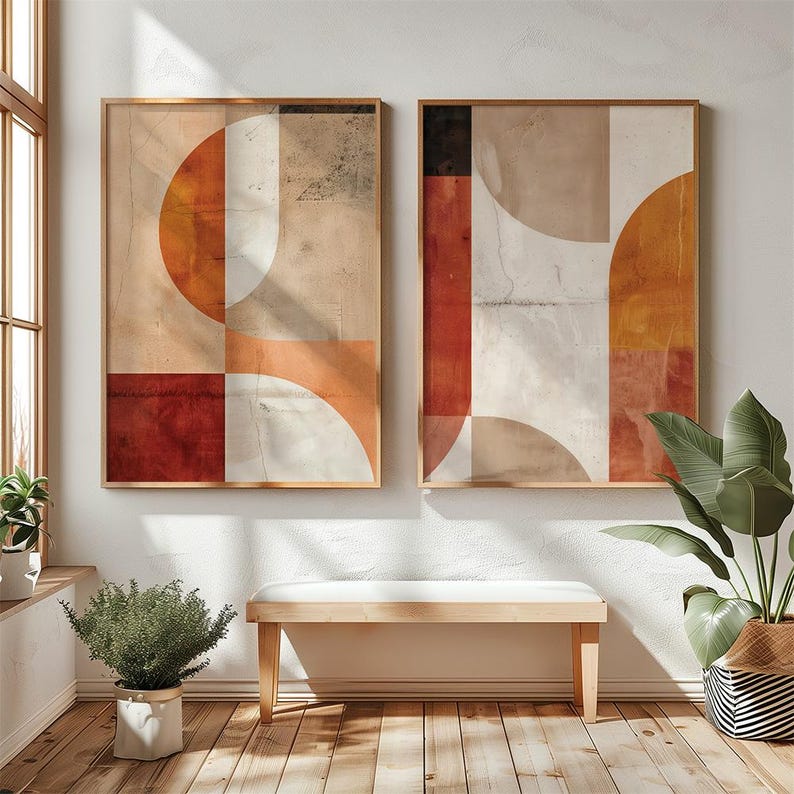 May include: Two framed abstract art prints. The artwork features geometric shapes in shades of red, orange, white, and beige. The frames are a light brown color. The prints are displayed above a wooden bench in a room with a window and plants.