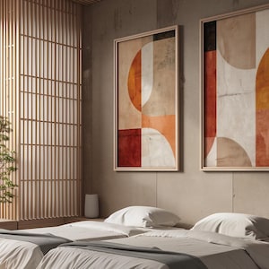 May include: Two framed abstract art prints with geometric shapes in shades of orange, red, and beige. The artwork is displayed on a neutral-colored wall above a bed with white bedding. The room features a wooden screen and a minimalist aesthetic.