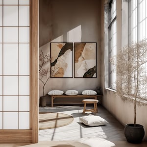 May include: A minimalist living room with a wooden bench and two white cushions, a round wooden stool, and a white throw pillow. Two framed abstract art prints with brown, white, and green colors hang on the wall. A small tree with brown branches is in the corner of the room.