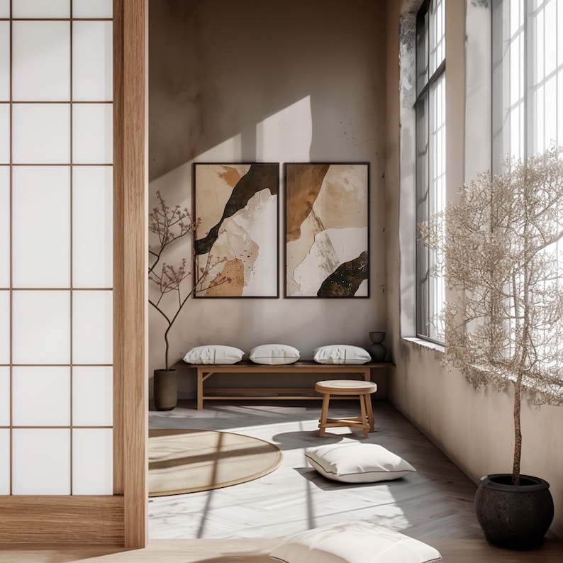 May include: A minimalist living room with a wooden bench and two white cushions, a round wooden stool, and a white throw pillow. Two framed abstract art prints with brown, white, and green colors hang on the wall. A small tree with brown branches is in the corner of the room.