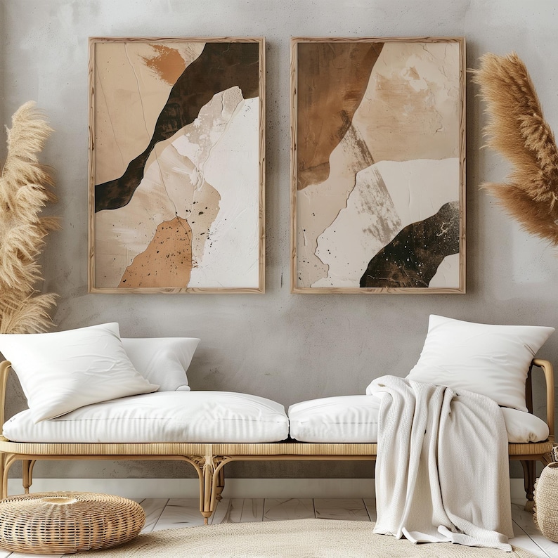 May include: Two abstract art prints in wooden frames with brown, beige, and black abstract shapes. The prints are hanging on a light gray wall above a white wicker bench with white cushions and a white throw blanket.