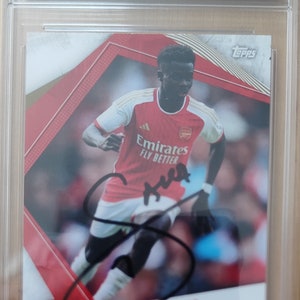 Puede incluir: Una tarjeta de deporte clasificada con una foto de Bukayo Saka, un futbolista profesional del Arsenal FC. La tarjeta está firmada por Saka y tiene una calificación de 9. La tarjeta es de la serie Topps-Signed 2023/24.