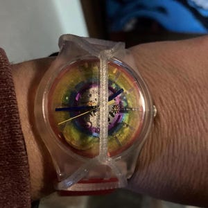 May include: A Swatch wristwatch featuring a transparent plastic case and a unique cage-like overlay. The dial displays a vibrant rainbow gradient with visible internal mechanical gears, complemented by thin blue and yellow hands for timekeeping.
