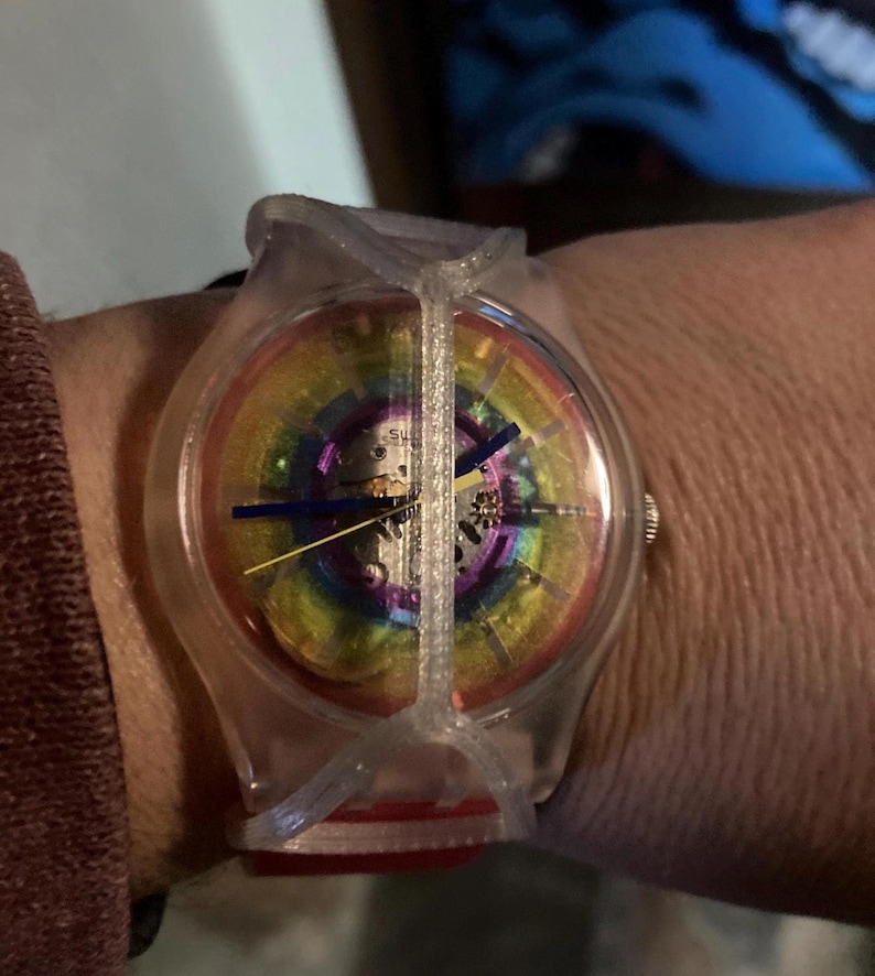 May include: A Swatch wristwatch featuring a transparent plastic case and a unique cage-like overlay. The dial displays a vibrant rainbow gradient with visible internal mechanical gears, complemented by thin blue and yellow hands for timekeeping.