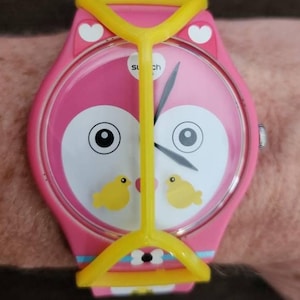 May include: A pink wristwatch featuring a stylized owl face on the dial with two small yellow birds, white heart accents, and a bright yellow plastic frame overlay. The watch has a pink strap with blue and white details and black clock hands.