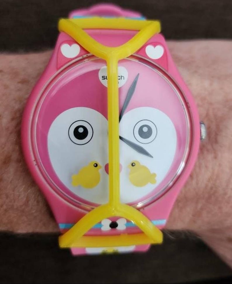 May include: A pink wristwatch featuring a stylized owl face on the dial with two small yellow birds, white heart accents, and a bright yellow plastic frame overlay. The watch has a pink strap with blue and white details and black clock hands.