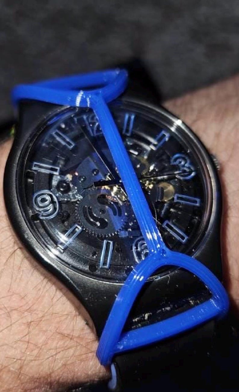 May include: A black wristwatch with a skeleton dial featuring blue hour markers and hands. A bright blue, Y-shaped plastic frame is attached over the watch face, extending from the top to the bottom of the watch case, securing over the black strap.
