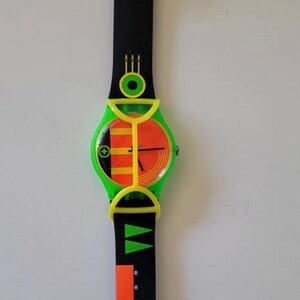 May include: A wristwatch featuring a black strap with orange and green geometric patterns, a bright green plastic guard over the round face, and a dial displaying orange and yellow vertical stripes with black hands and a small circular green accent.