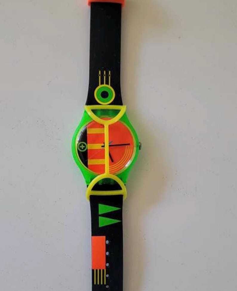 May include: A wristwatch featuring a black strap with orange and green geometric patterns, a bright green plastic guard over the round face, and a dial displaying orange and yellow vertical stripes with black hands and a small circular green accent.