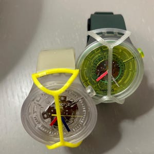May include: Two Swatch wristwatches with exposed mechanical movements. One features a clear case, a yellow protective frame, and red and blue hands. The other has a dark green strap, a clear case, a lime green dial, and a white protective frame.