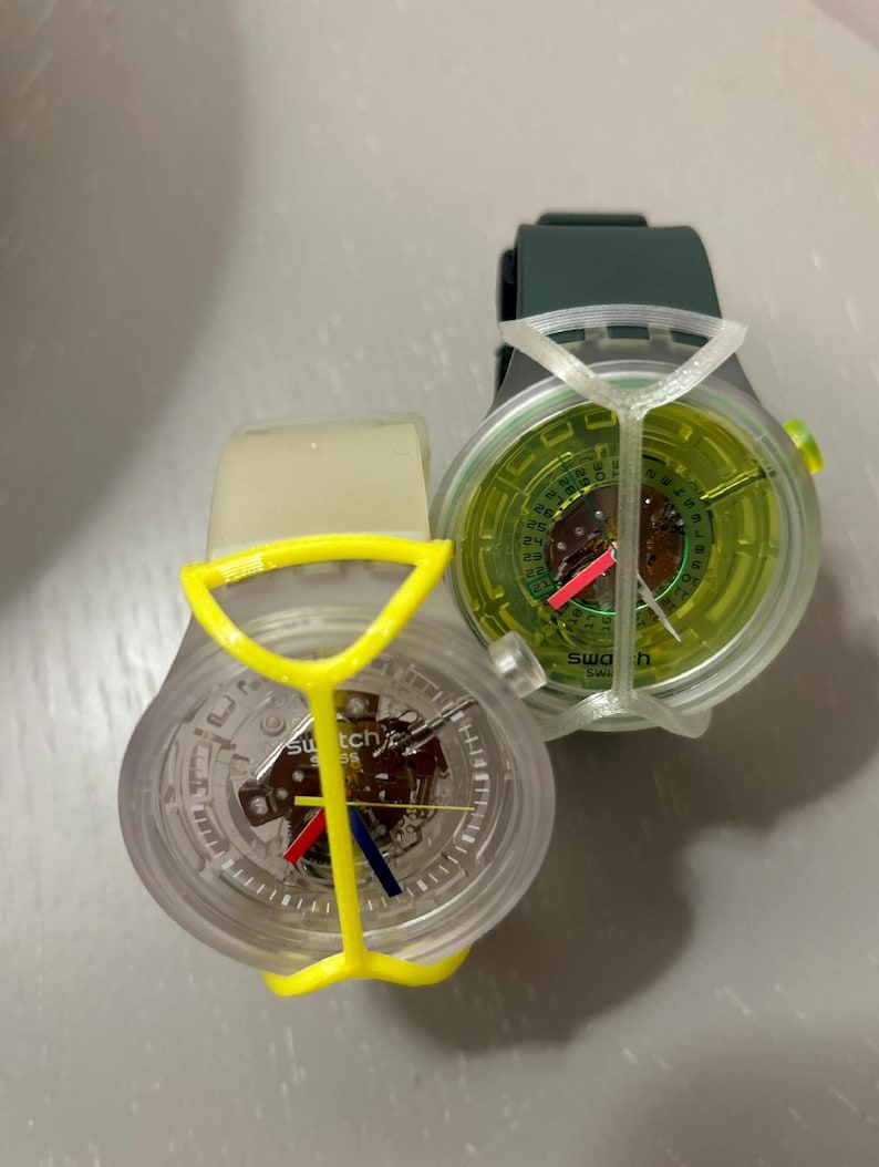 May include: Two Swatch wristwatches with exposed mechanical movements. One features a clear case, a yellow protective frame, and red and blue hands. The other has a dark green strap, a clear case, a lime green dial, and a white protective frame.