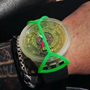 May include: A wristwatch with a translucent yellow dial, a black silicone strap, and a bright green plastic frame overlay. The watch face features visible internal gears, a date wheel with numbers, and the text SWATCH printed on the lower dial.