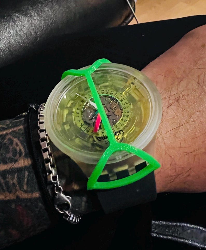 May include: A wristwatch with a translucent yellow dial, a black silicone strap, and a bright green plastic frame overlay. The watch face features visible internal gears, a date wheel with numbers, and the text SWATCH printed on the lower dial.