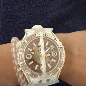May include: A white wristwatch with a transparent case and a skeleton dial featuring the numbers 3, 6, and 9. The watch has a rotating bezel with numerical markings and a protective white frame. A translucent beaded bracelet is worn alongside it.