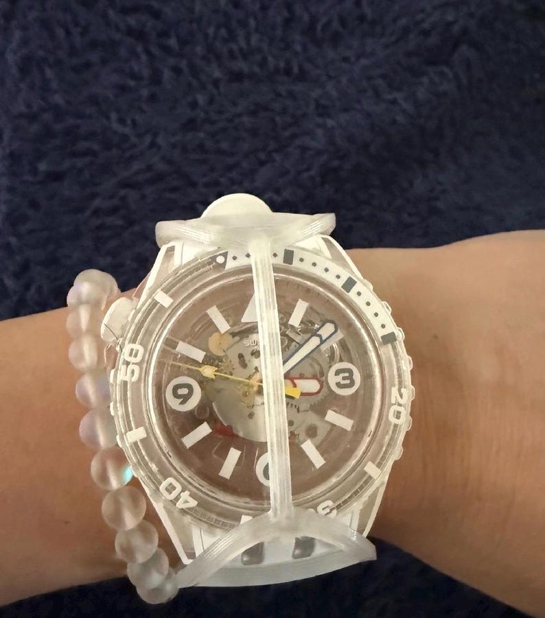 May include: A white wristwatch with a transparent case and a skeleton dial featuring the numbers 3, 6, and 9. The watch has a rotating bezel with numerical markings and a protective white frame. A translucent beaded bracelet is worn alongside it.