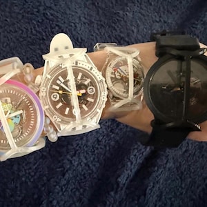 May include: Four Swatch wristwatches featuring protective white silicone cages. The first watch has a rainbow-colored bezel, the second and third are clear with visible internal mechanisms, and the fourth is a solid black watch with a black strap.