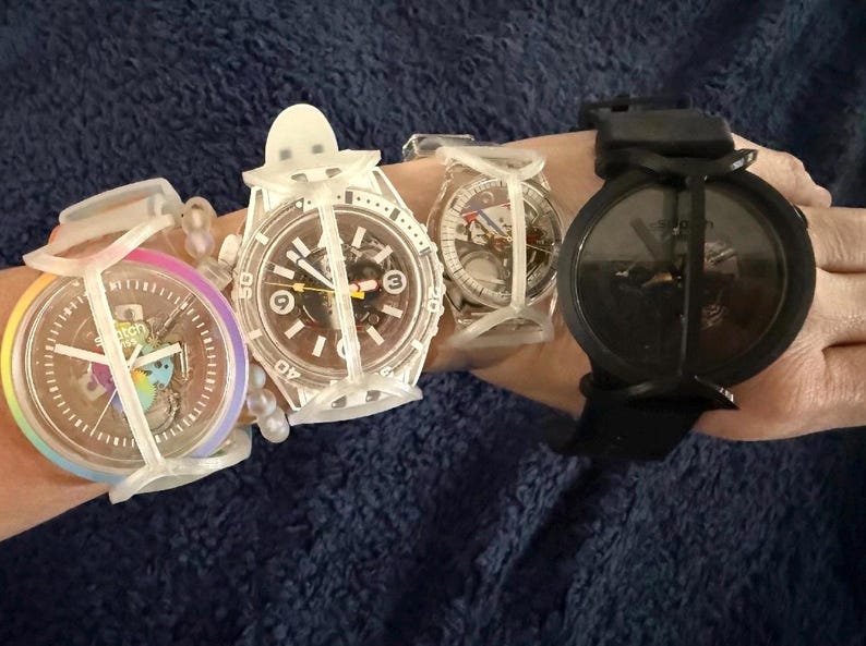 May include: Four Swatch wristwatches featuring protective white silicone cages. The first watch has a rainbow-colored bezel, the second and third are clear with visible internal mechanisms, and the fourth is a solid black watch with a black strap.