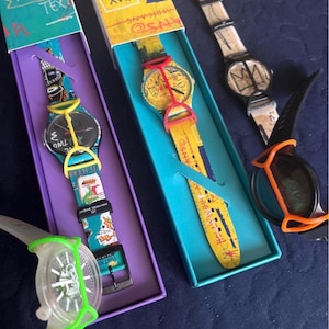 May include: Four wristwatches featuring artistic graphic designs and colorful silicone protective covers. Two watches are housed in rectangular boxes with purple and teal interiors. The watches display various abstract patterns, text, and illustrations.