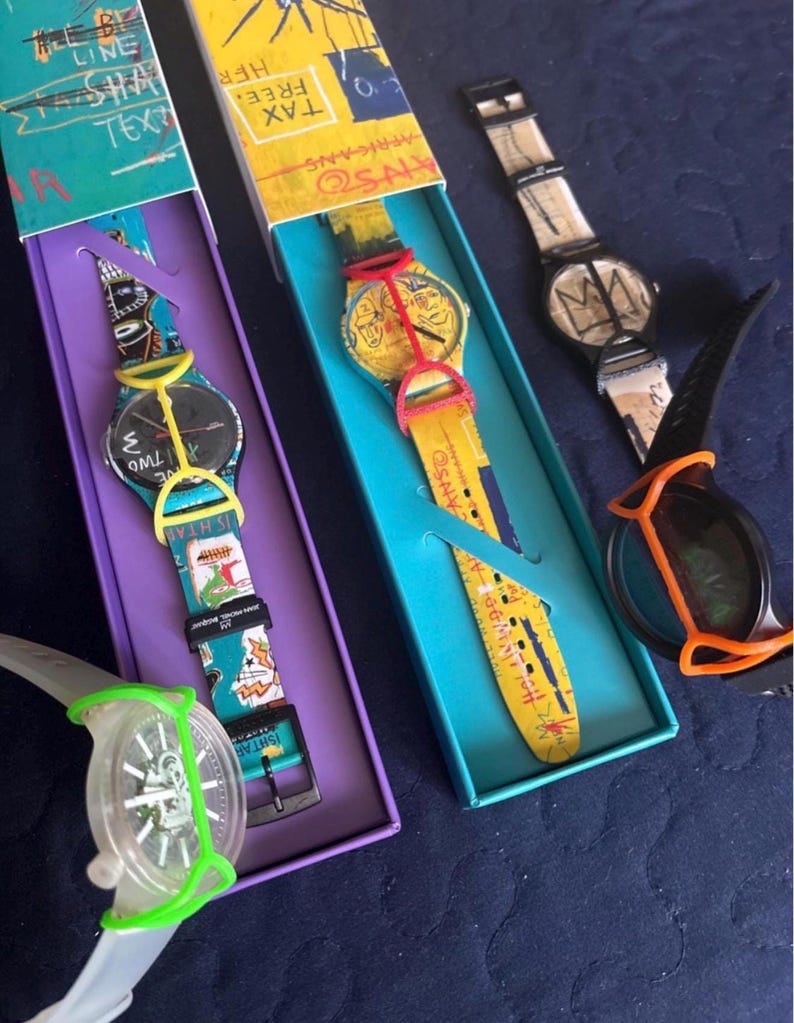 May include: Four wristwatches featuring artistic graphic designs and colorful silicone protective covers. Two watches are housed in rectangular boxes with purple and teal interiors. The watches display various abstract patterns, text, and illustrations.