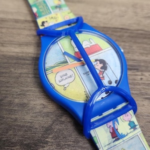 May include: A blue wristwatch featuring a Peanuts comic strip design on the watch face and strap. The dial displays the text "WING WALKING!" and "swatch SWISS". A blue plastic guard covers the watch face, and the strap shows various characters.