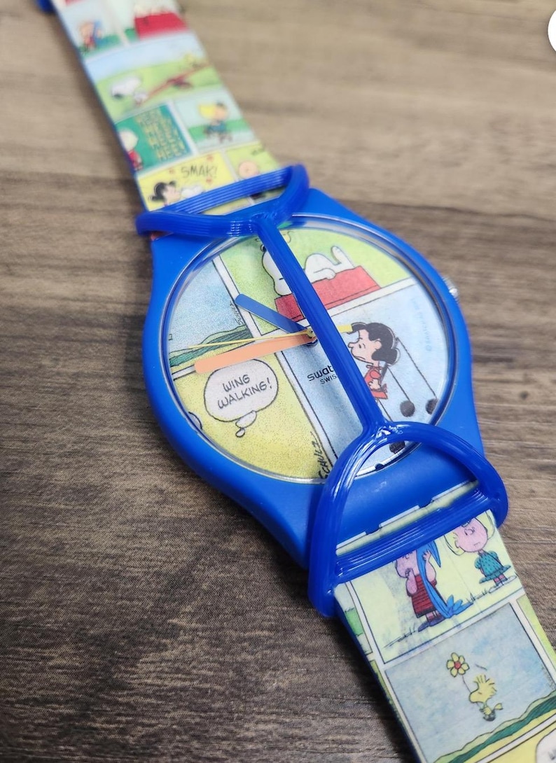 May include: A blue wristwatch featuring a Peanuts comic strip design on the watch face and strap. The dial displays the text "WING WALKING!" and "swatch SWISS". A blue plastic guard covers the watch face, and the strap shows various characters.