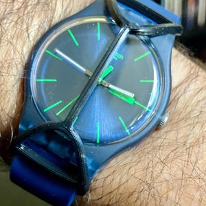 May include: A dark blue wristwatch featuring a circular face with bright green hour markers and hands. The watch has a dark blue silicone strap and a black plastic protective guard covering the crystal. The word Swatch is visible on the dial.
