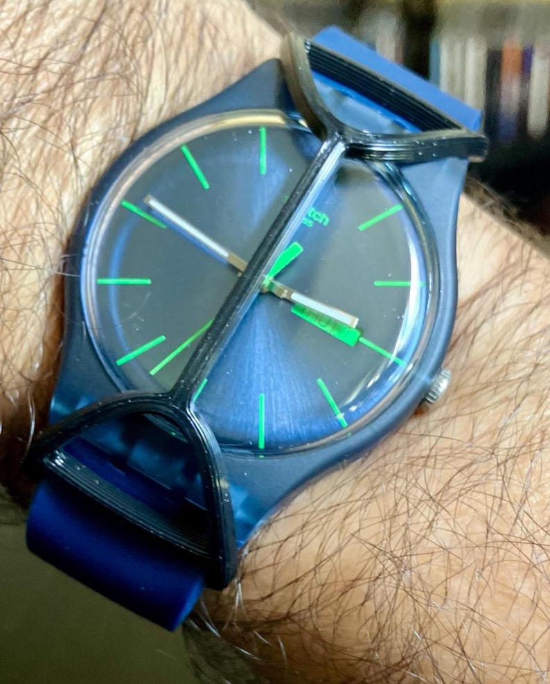May include: A dark blue wristwatch featuring a circular face with bright green hour markers and hands. The watch has a dark blue silicone strap and a black plastic protective guard covering the crystal. The word Swatch is visible on the dial.